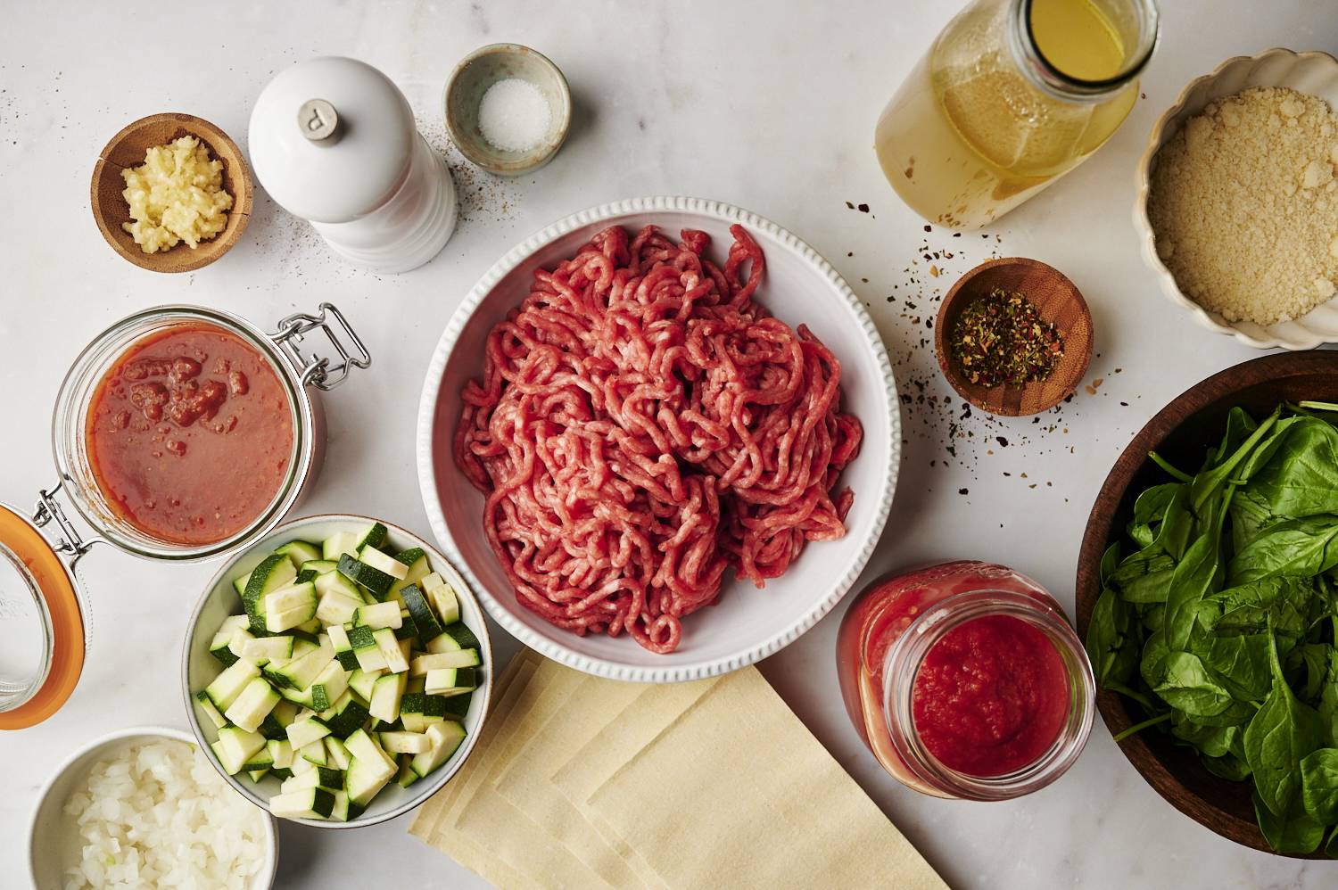 Ingredients for slow cooker lasagna soup, including ground meat, zucchini, spinach, lasagna noodles, tomato sauce, and seasonings, arranged on a white surface.