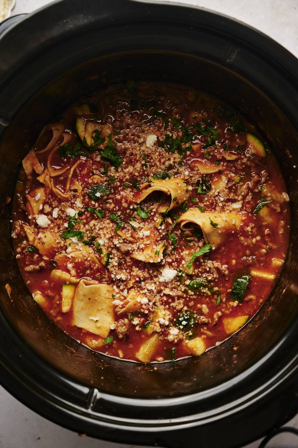 Slow cooker lasagna soup with ground meat, zucchini, pasta, and parmesan cheese, garnished with fresh parsley.