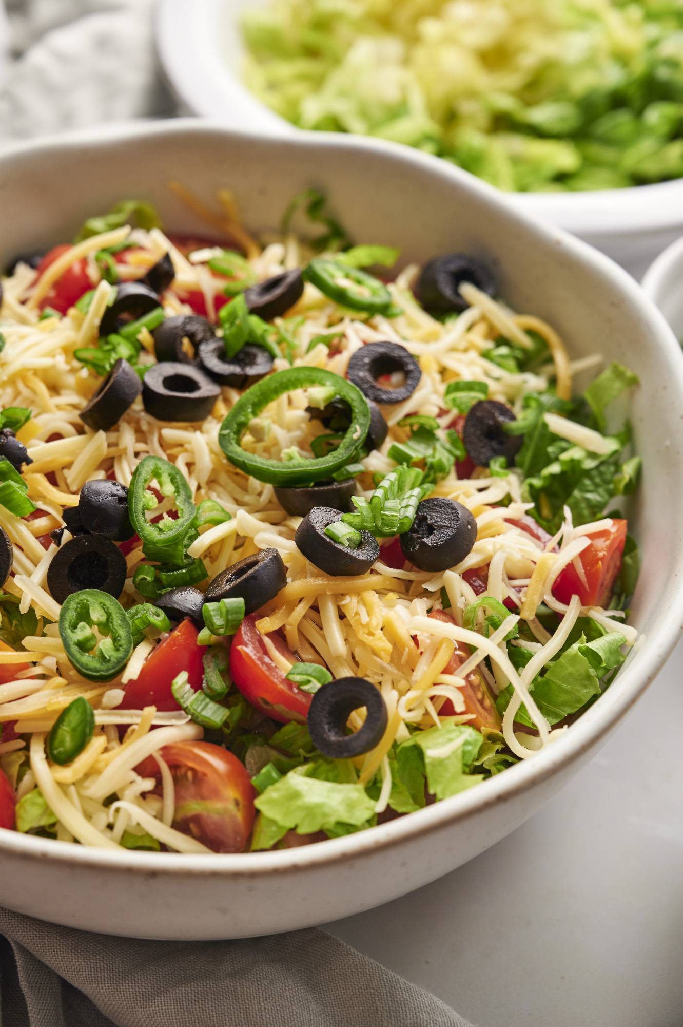 Bowl of healthy taco dip topped with lettuce, cheese, tomatoes, black olives, green onions, and jalapenos.