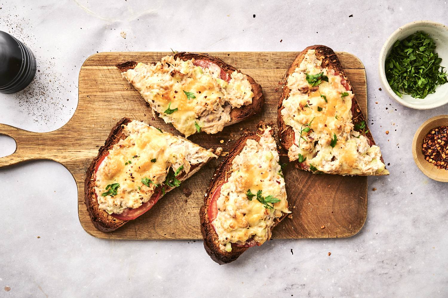 Tuna salad melts with sliced tomatoes, melted cheddar cheese, and parsley served on whole wheat bread on a wooden cutting board.