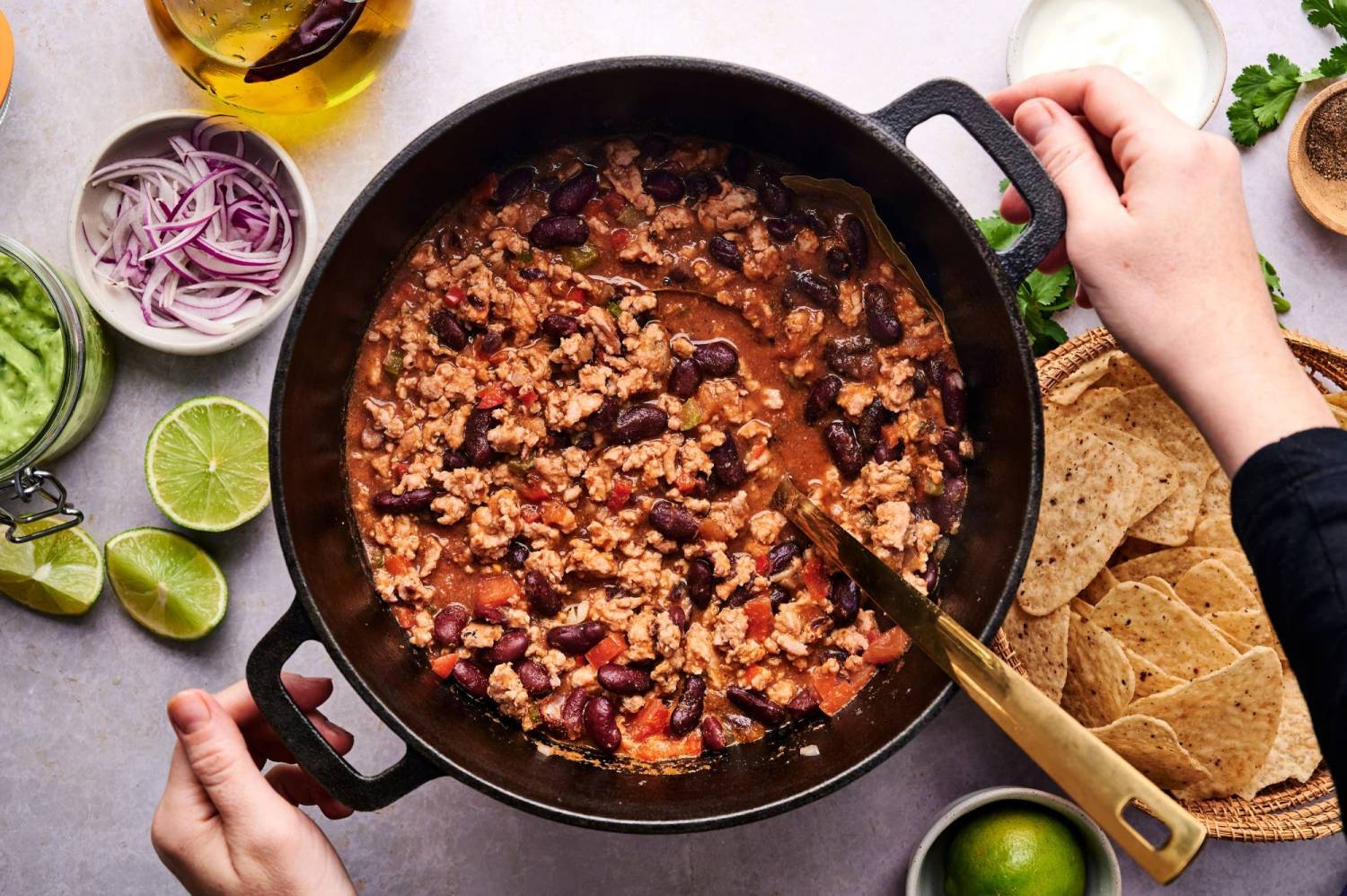 Pot of healthy turkey chili with ground turkey, kidney beans, tomatoes, and peppers with chips, lime, cilantro, cheese, and sour cream on the side.