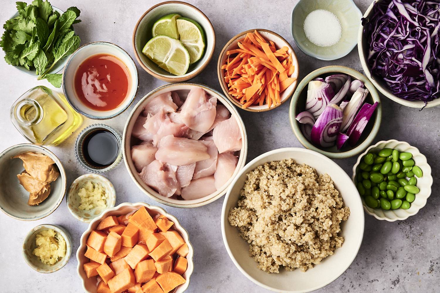 An array of fresh ingredients for a meal, including fish, quinoa, vegetables, herbs, and sauces, arranged in small bowls on a countertop.