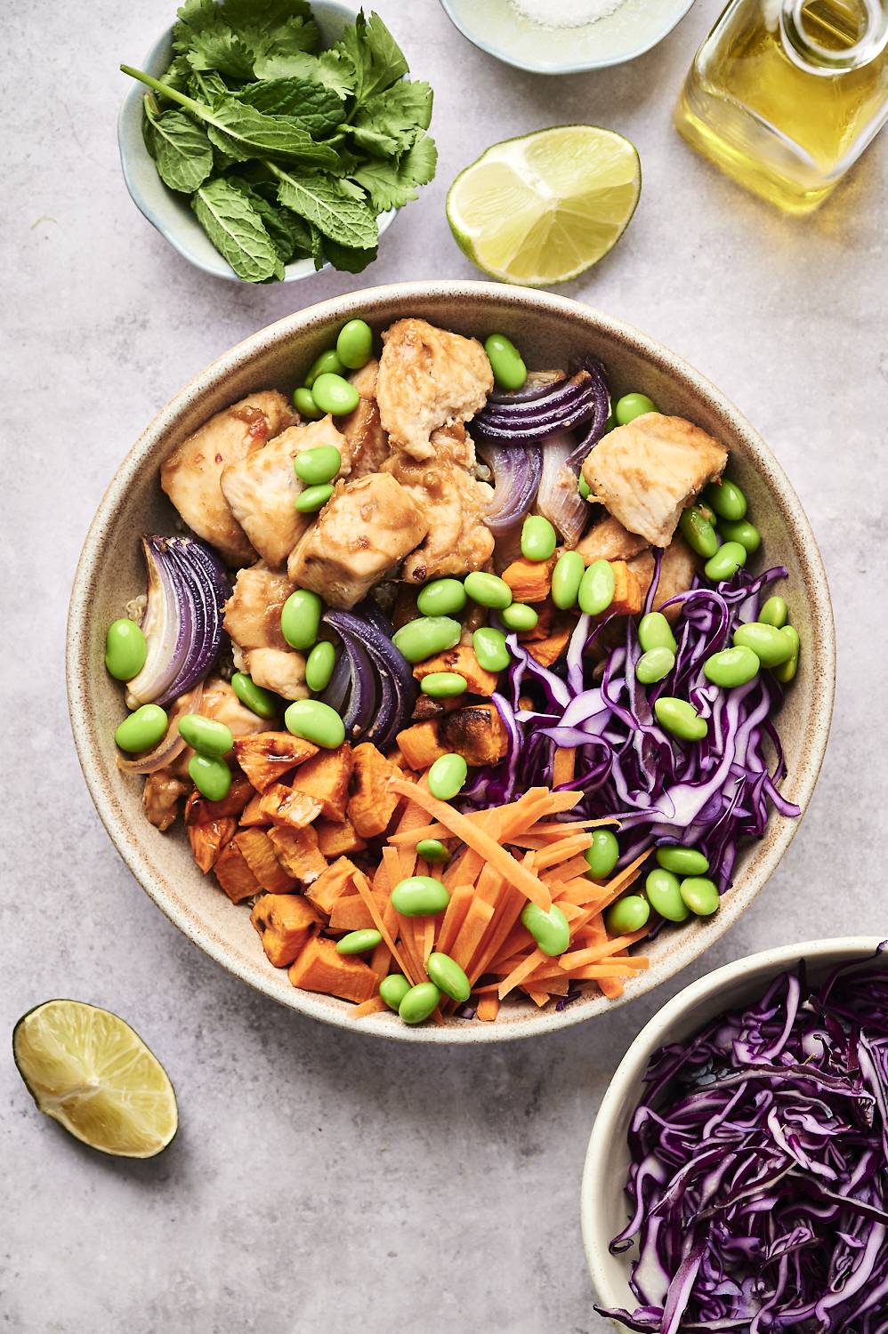 A vibrant bowl of chicken, edamame, sweet potatoes, carrots, and purple cabbage, garnished with mint and lime.