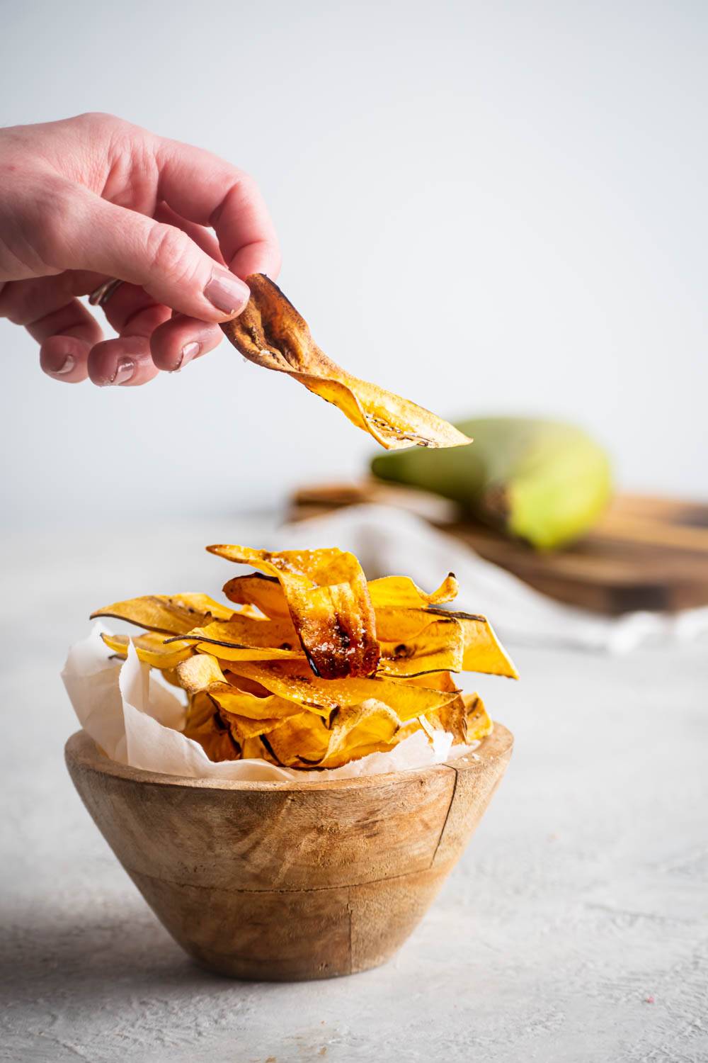Hand holding a crispy homemade plantain chip above a rustic wooden bowl filled with golden chips, with a green plantain in the background.