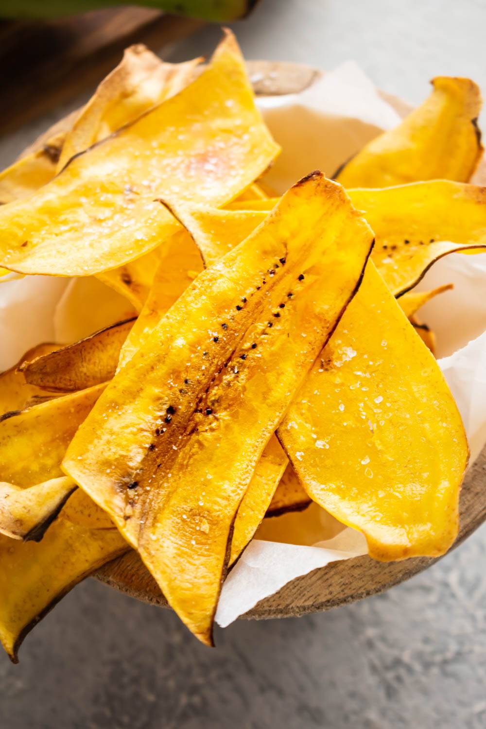 Close-up of crispy homemade plantain chips sprinkled with sea salt in a wooden bowl lined with parchment paper.
