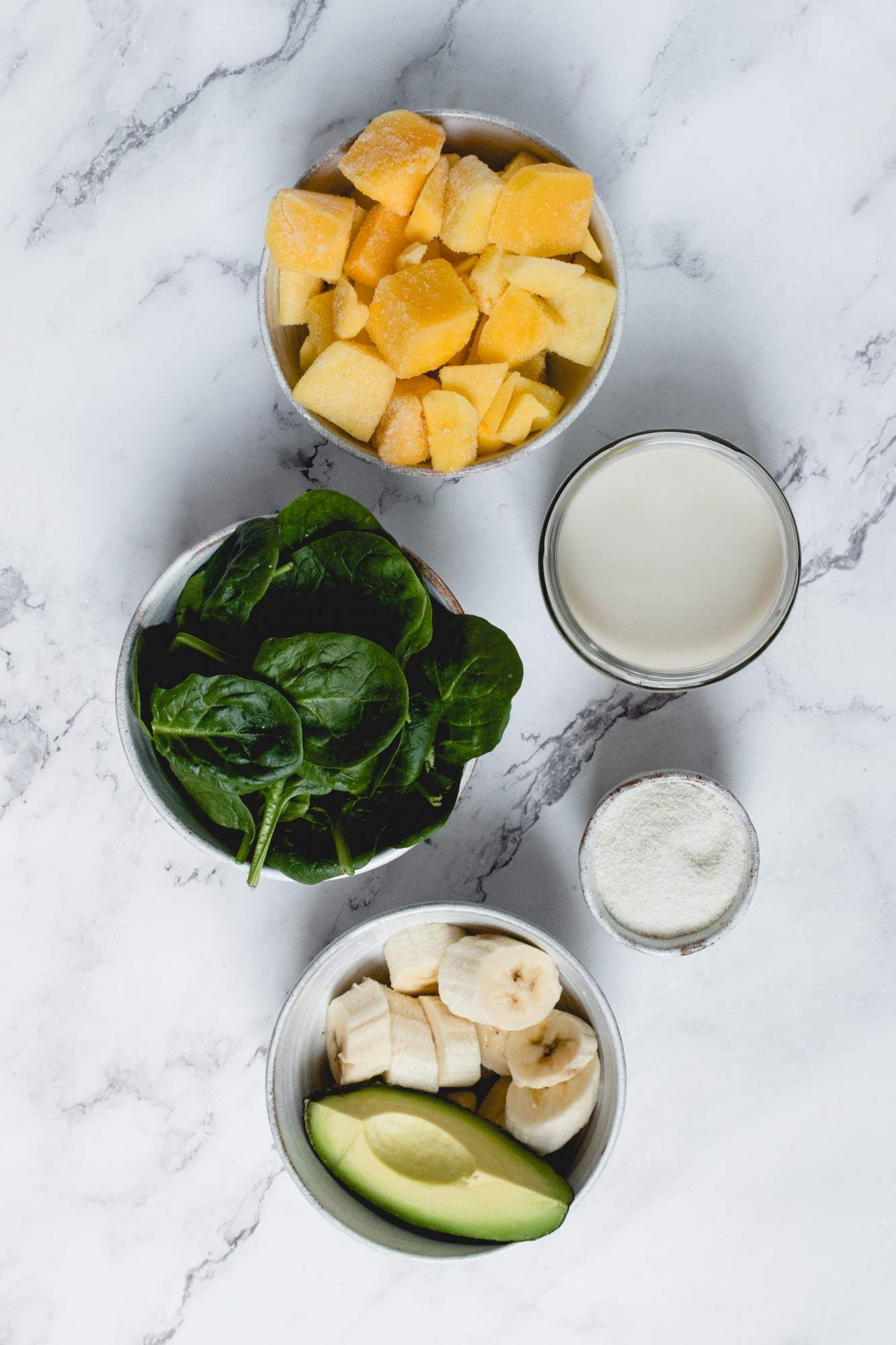 Ingredients for a mango and spinach smoothie including spinach leaves, avocado, banana, frozen mango, and protein powder.