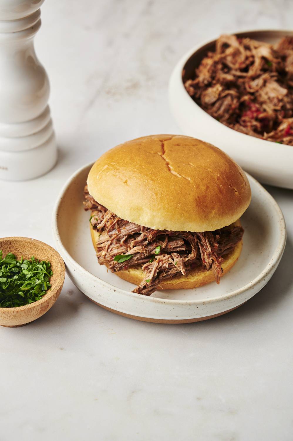 A shredded beef sandwich on a golden bun, served on a rustic ceramic plate with a small wooden bowl of chopped parsley nearby and a bowl of extra shredded beef in the background.