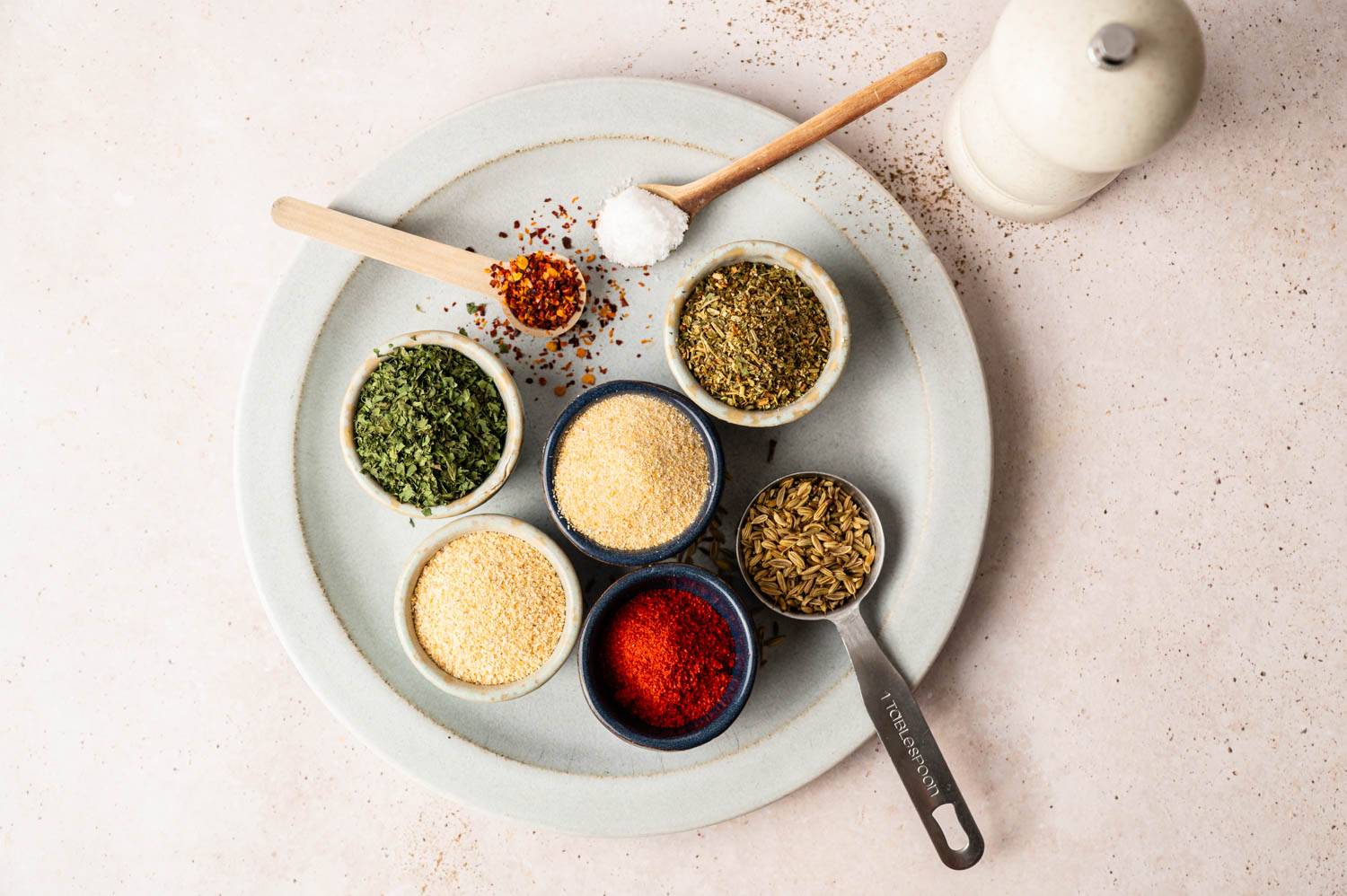 A round plate holds bowls of various spices, including dried herbs, garlic powder, paprika, and fennel, with small wooden spoons and a salt shaker nearby.