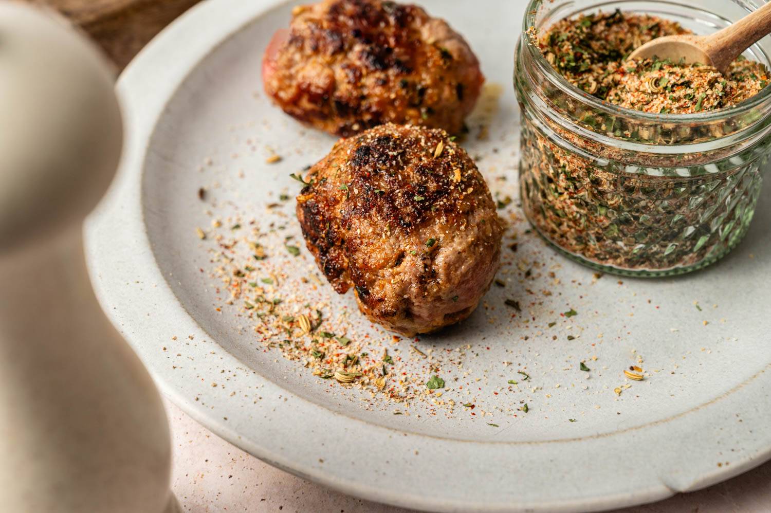 Two browned meatballs on a white plate, sprinkled with herbs. A jar of seasoning with a small wooden spoon is beside them, creating a savory, homemade feel.