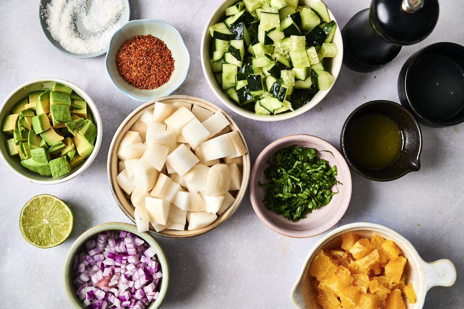 An assortment of fresh ingredients for a salad: chopped avocado, cucumber, jicama, red onion, cilantro, orange, spices, and lime.
