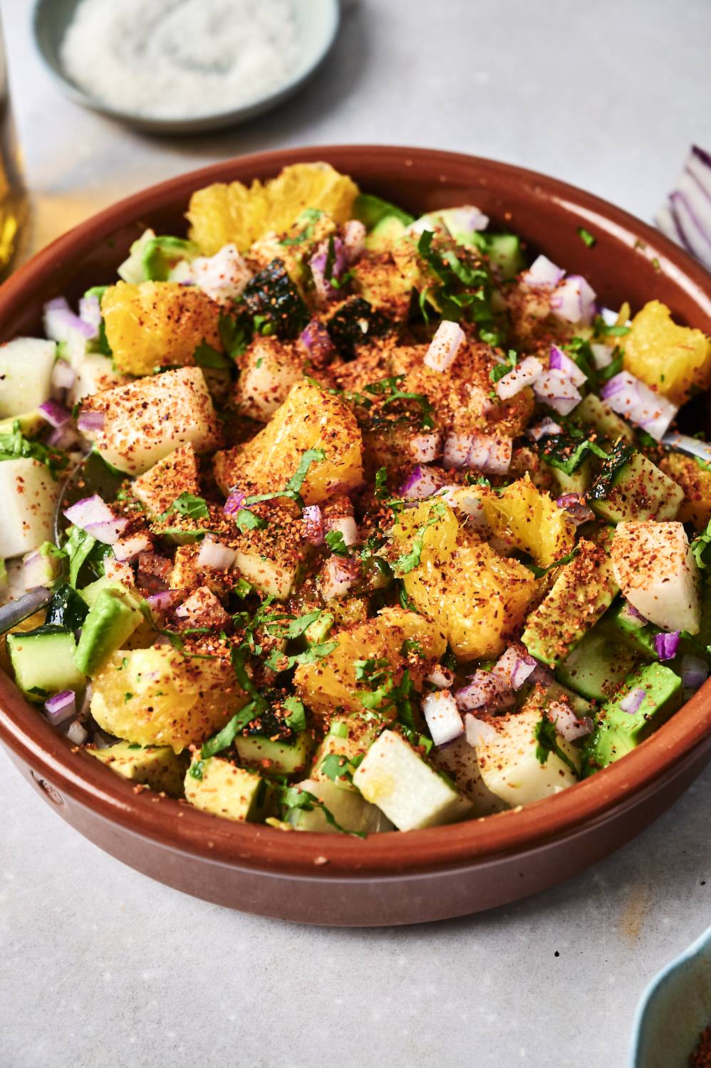 A colorful salad in a brown bowl, topped with croutons, orange slices, and spices, surrounded by fresh vegetables and herbs.