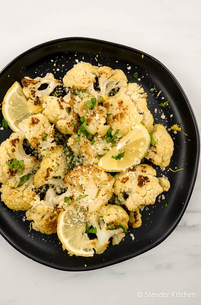 Roasted cauliflower on a black plate with  lemons, Parmesan cheese, and garlic.