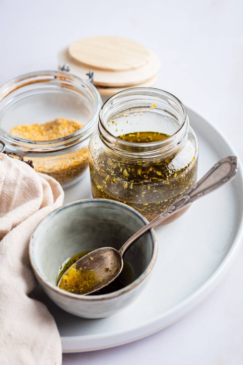 A glass jar of golden oil with herbs, accompanied by a small ceramic bowl with a spoon, sits on a round tray, creating a rustic, inviting scene.