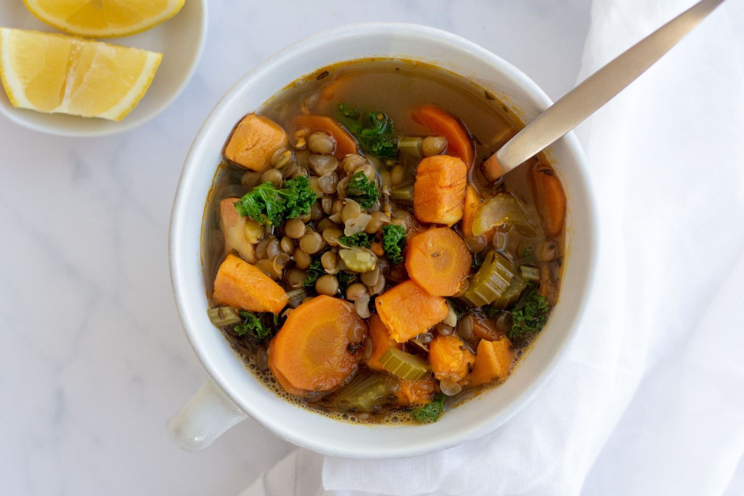 Lentil and vegetable soup in a white bowl with carrots, sweet potato, kale, lentils, and celery with a metal spoon and lemon slices.