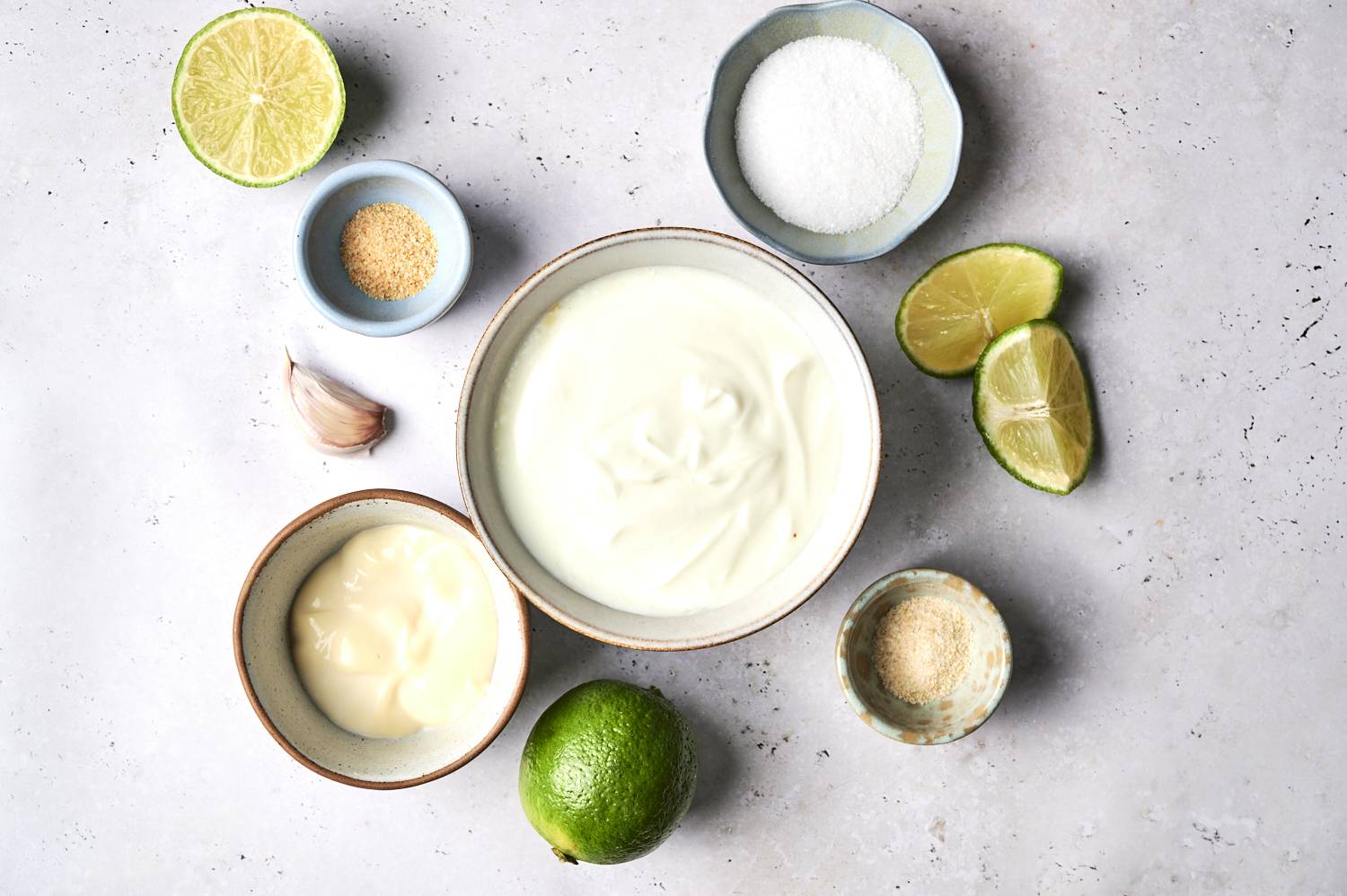 An array of ingredients on a light surface, including lime halves, yogurt, garlic, sugar, and spices in small bowls.
