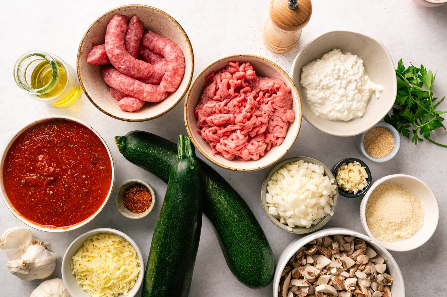 A variety of fresh ingredients for cooking: sausages, minced meat, cottage cheese, zucchinis, tomato sauce, herbs, chopped onions, garlic, and mushrooms.