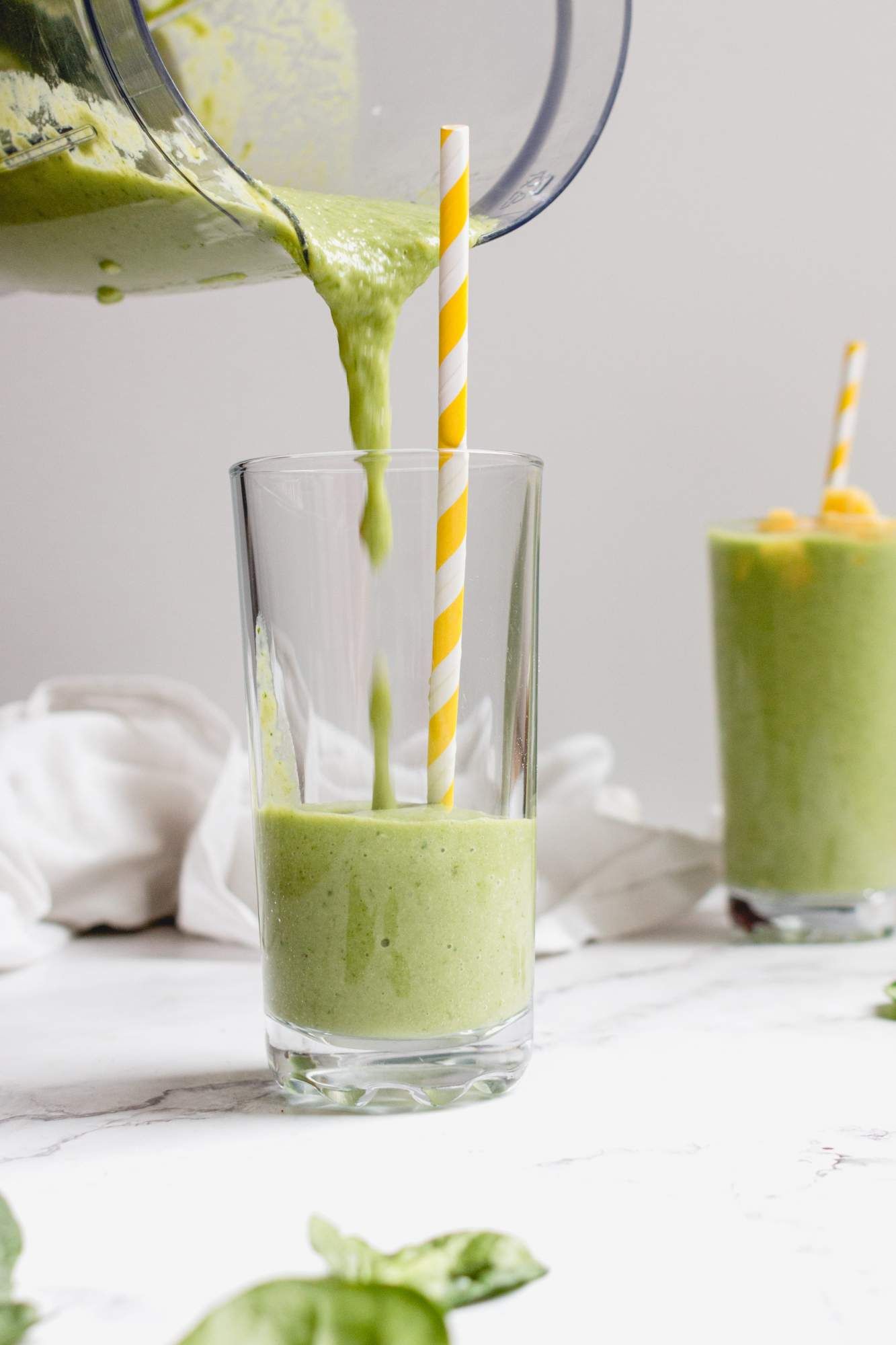 Mango green smoothie with spinach being poured from a blender into a glass with a yellow straw.