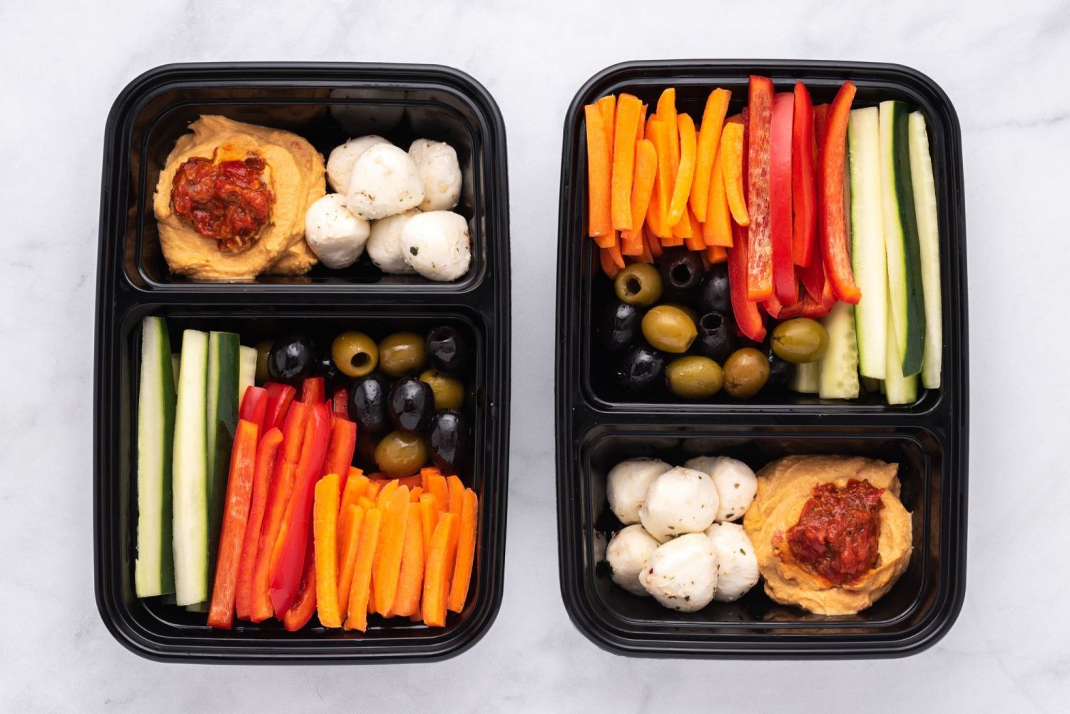 Two snack boxes packed with hummus, olives, vegetables, and cheese.