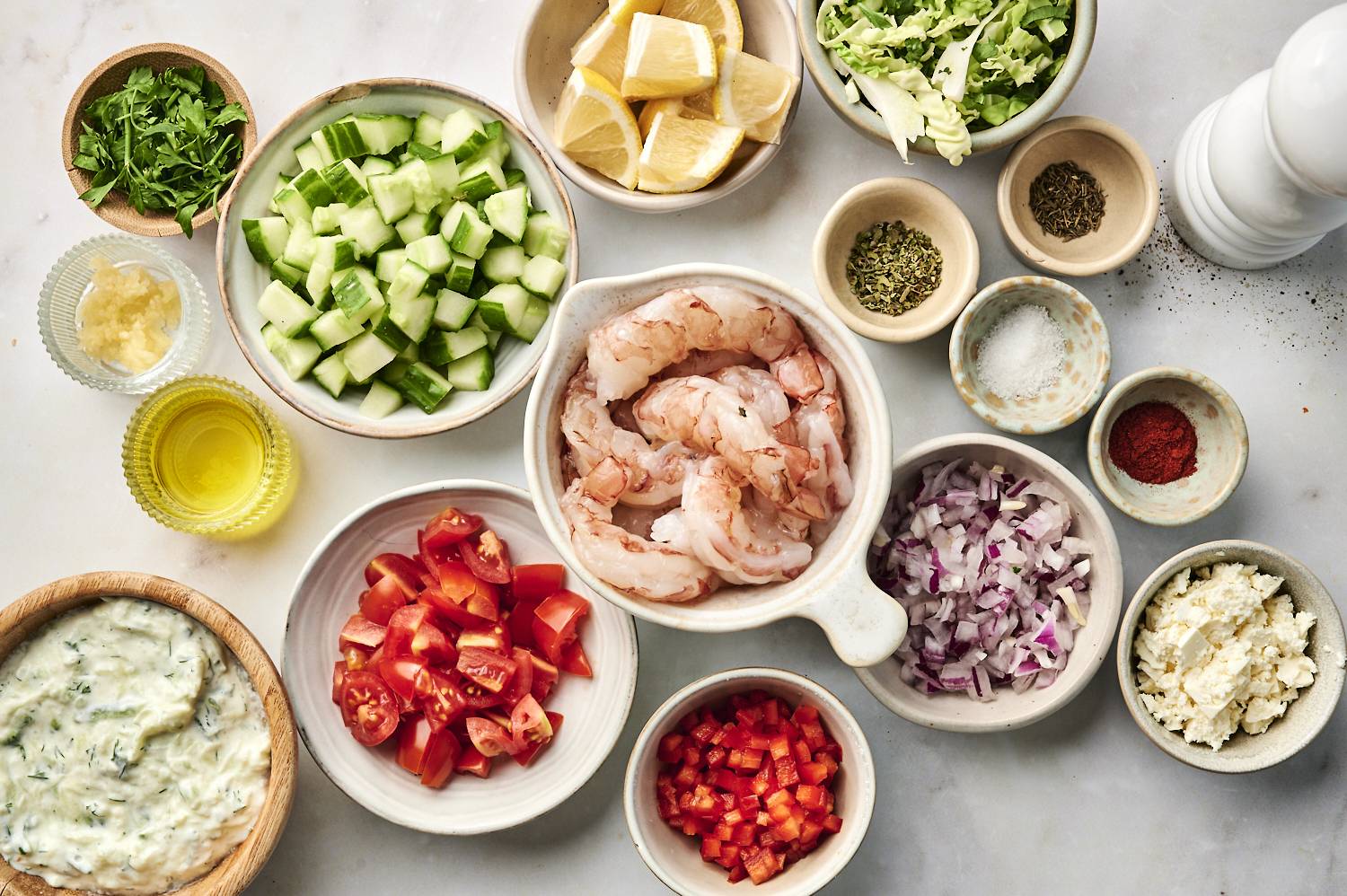 Ingredients for Mediterranean shrimp bowls, including raw shrimp, cucumbers, tomatoes, red onion, red bell peppers, lemon wedges, feta cheese, tzatziki, olive oil, garlic, and spices, arranged in bowls.