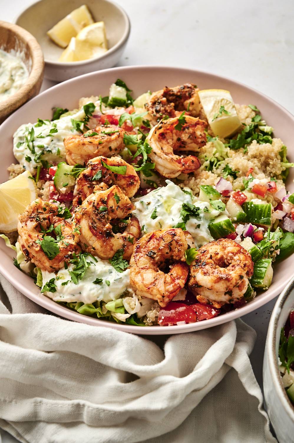 Close-up of Mediterranean shrimp bowls with grilled shrimp, quinoa, tzatziki sauce, fresh vegetables, and lemon wedges, garnished with parsley.