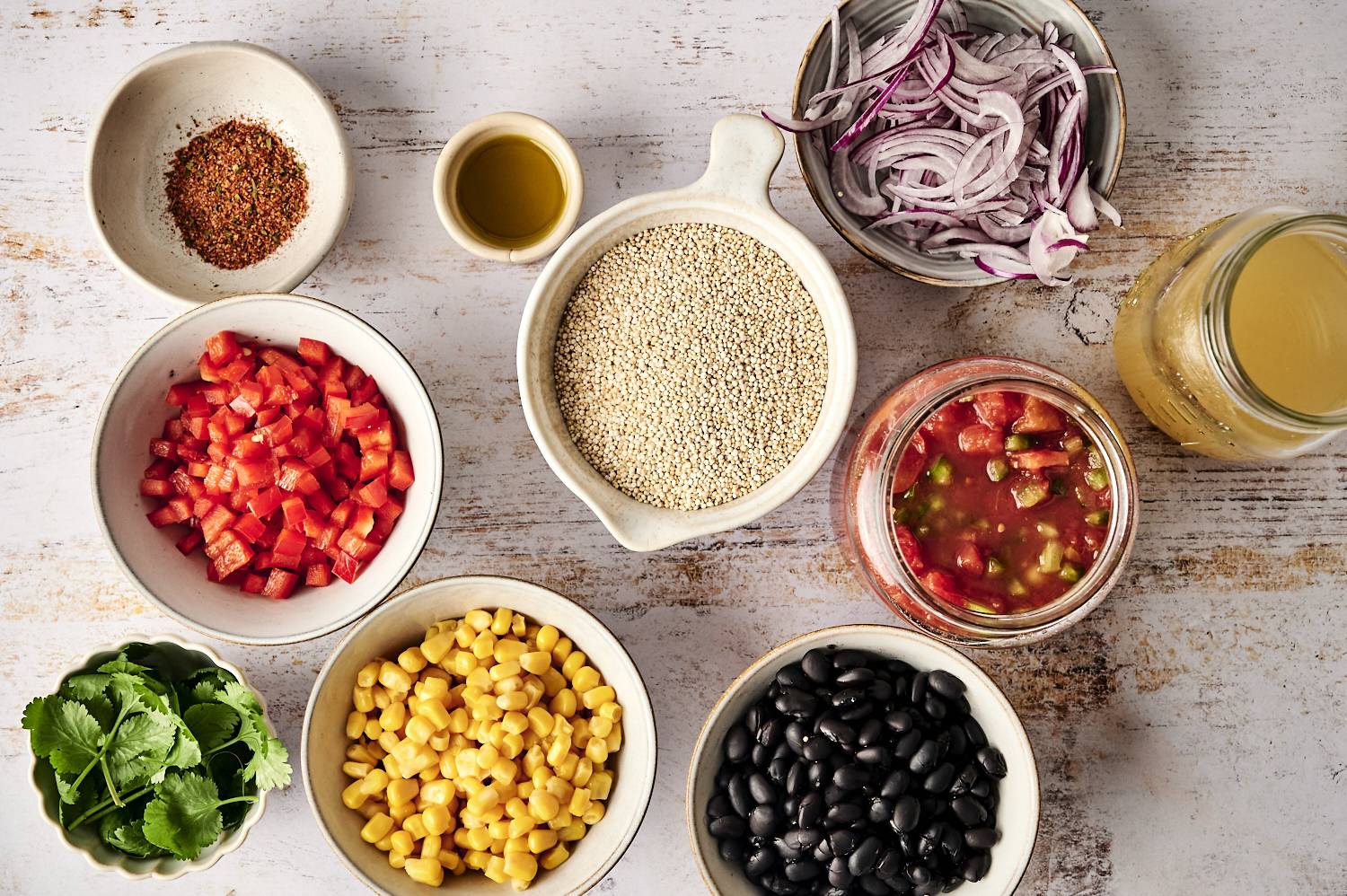 Ingredients for Mexican quinoa, including quinoa, black beans, corn, diced tomatoes, red onions, cilantro, and spices arranged on a wooden surface.