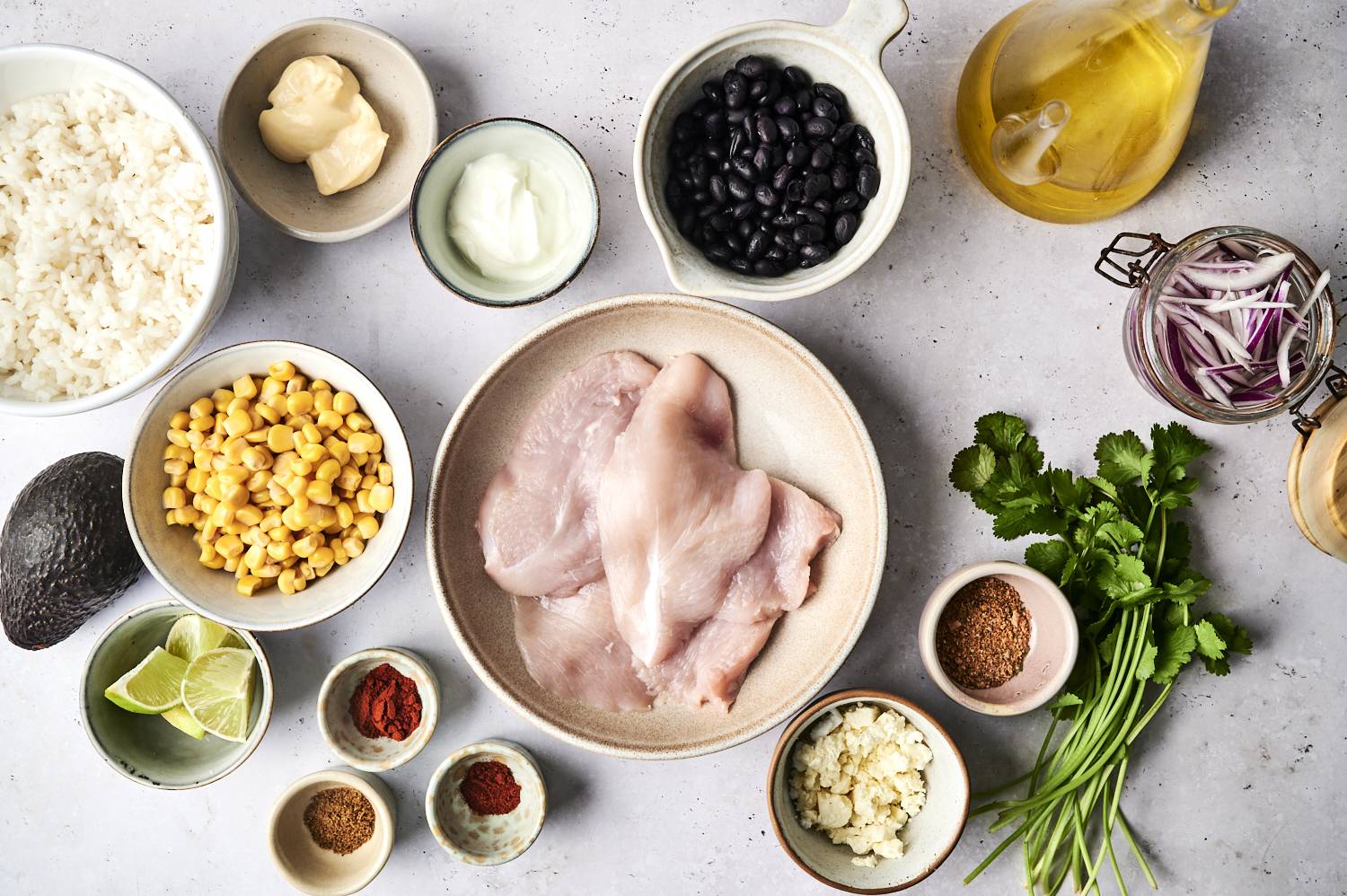 A top-down view of various ingredients: chicken fillets, black beans, corn, rice, spices, avocado, lime, and cilantro on a gray surface.