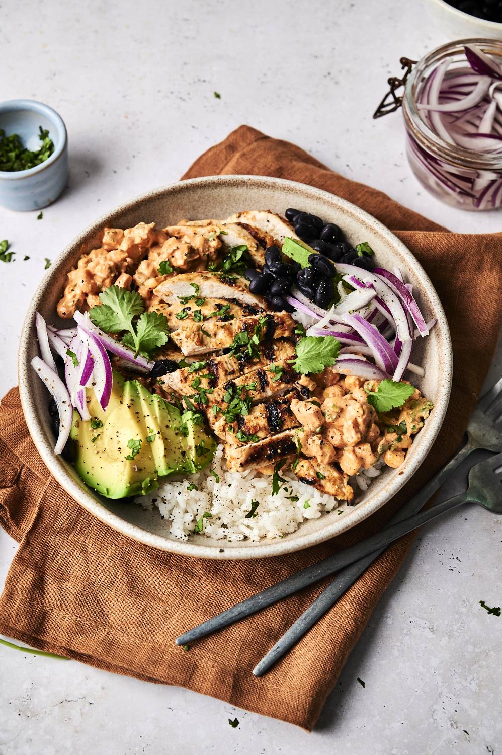 A vibrant bowl of grilled chicken, black beans, avocado, rice, and garnished with cilantro and red onion on a rustic cloth.