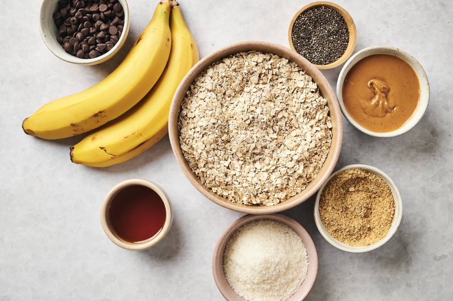 A flat lay of ripe bananas, bowls of oats, peanut butter, chocolate chips, chia seeds, and sweeteners on a light background.