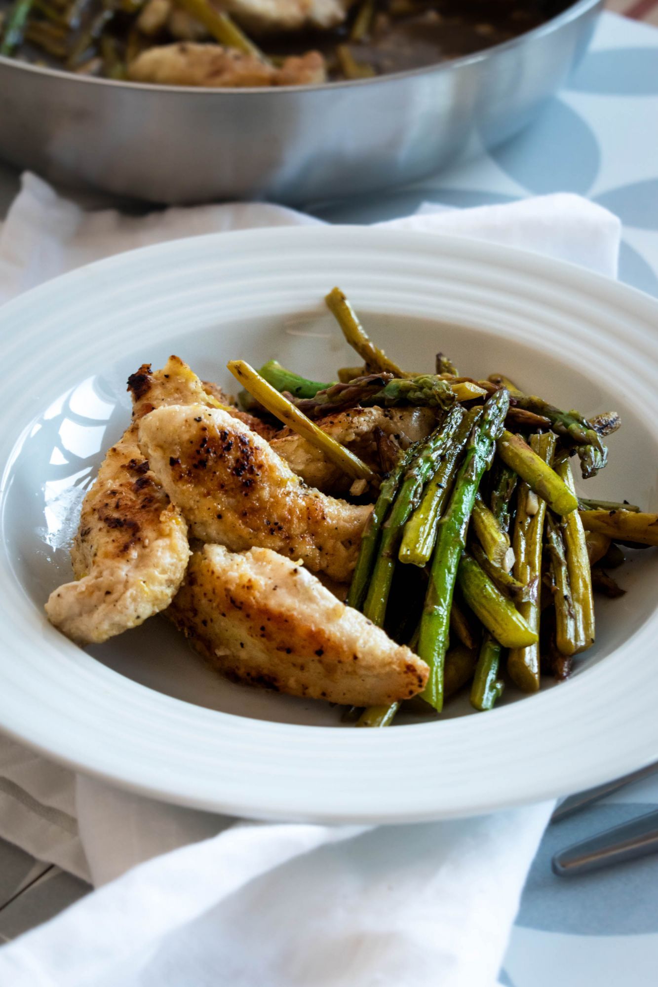 Lemon chicken and asparagus with a garlic pan sauce and crispy chicken served on a plate with a skillet in the background.