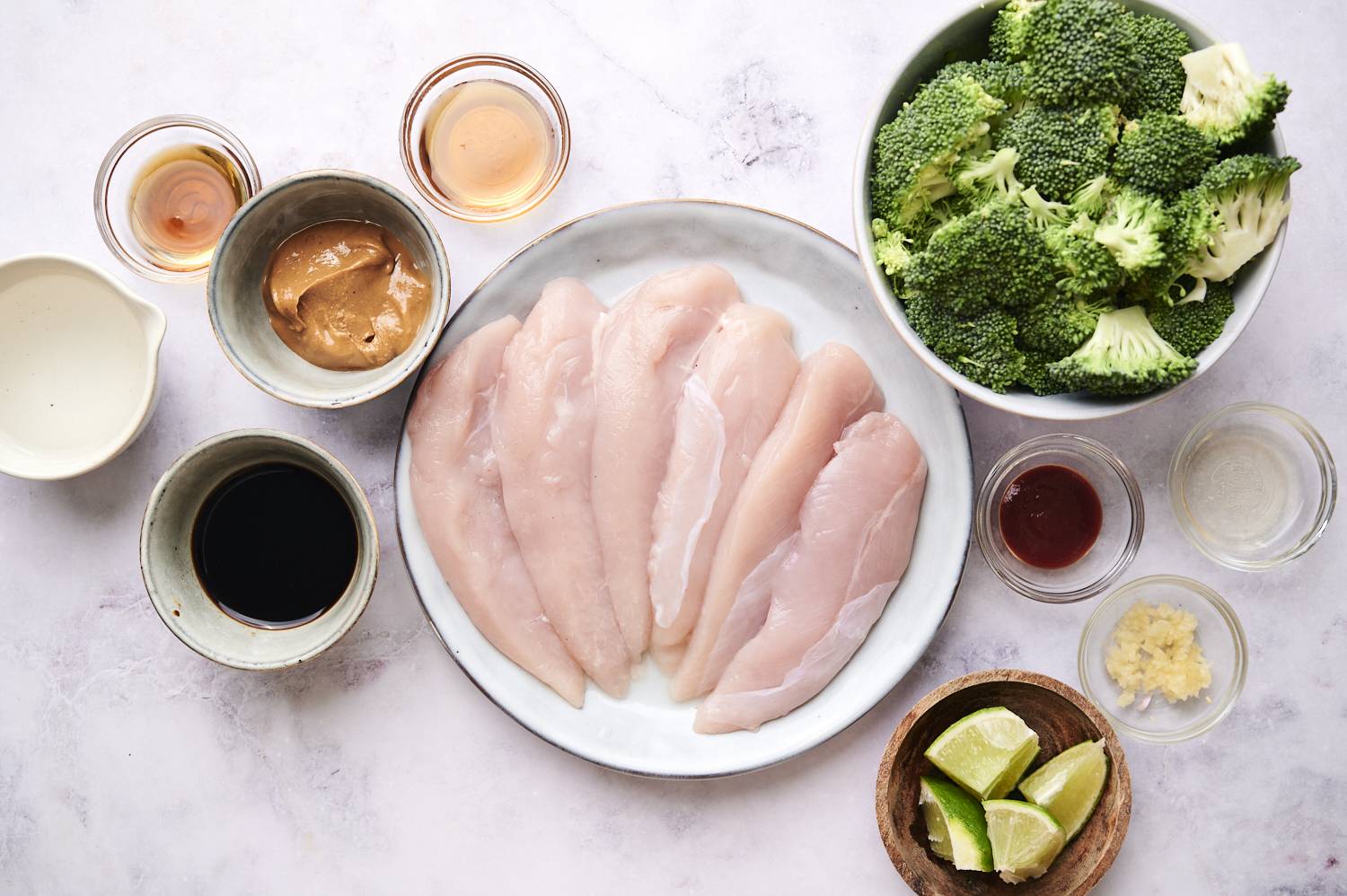 A hand stirring chicken marinade in a glass bowl, with raw chicken fillets on a plate nearby against a pale background.