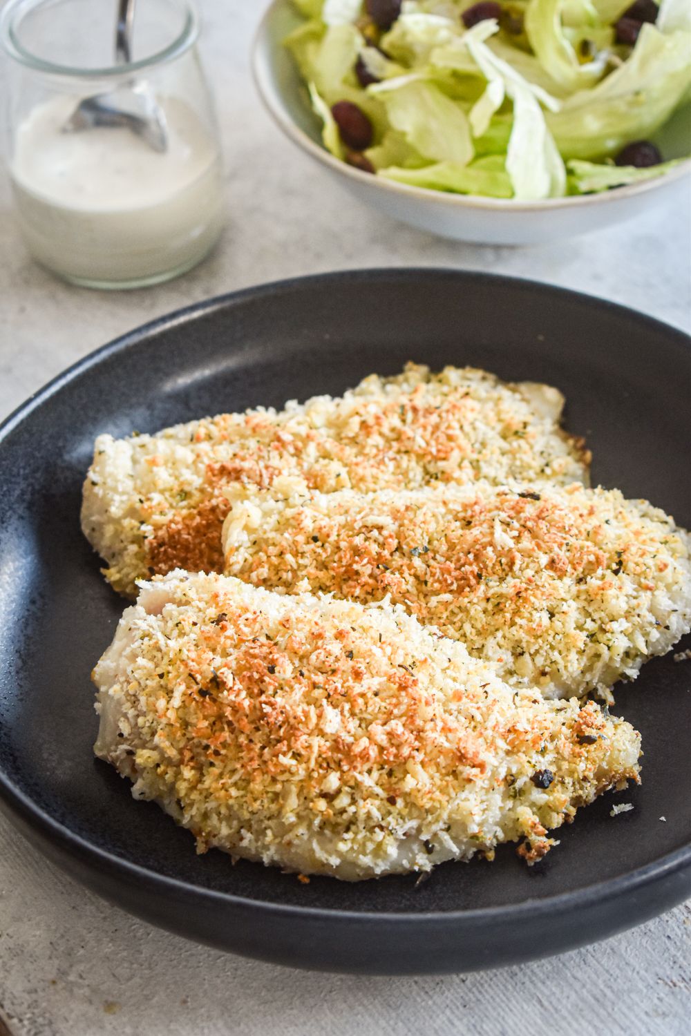 Tilapia crusted with Parmesan cheese and panko breadcrumbs on a black plate with salad on the side.