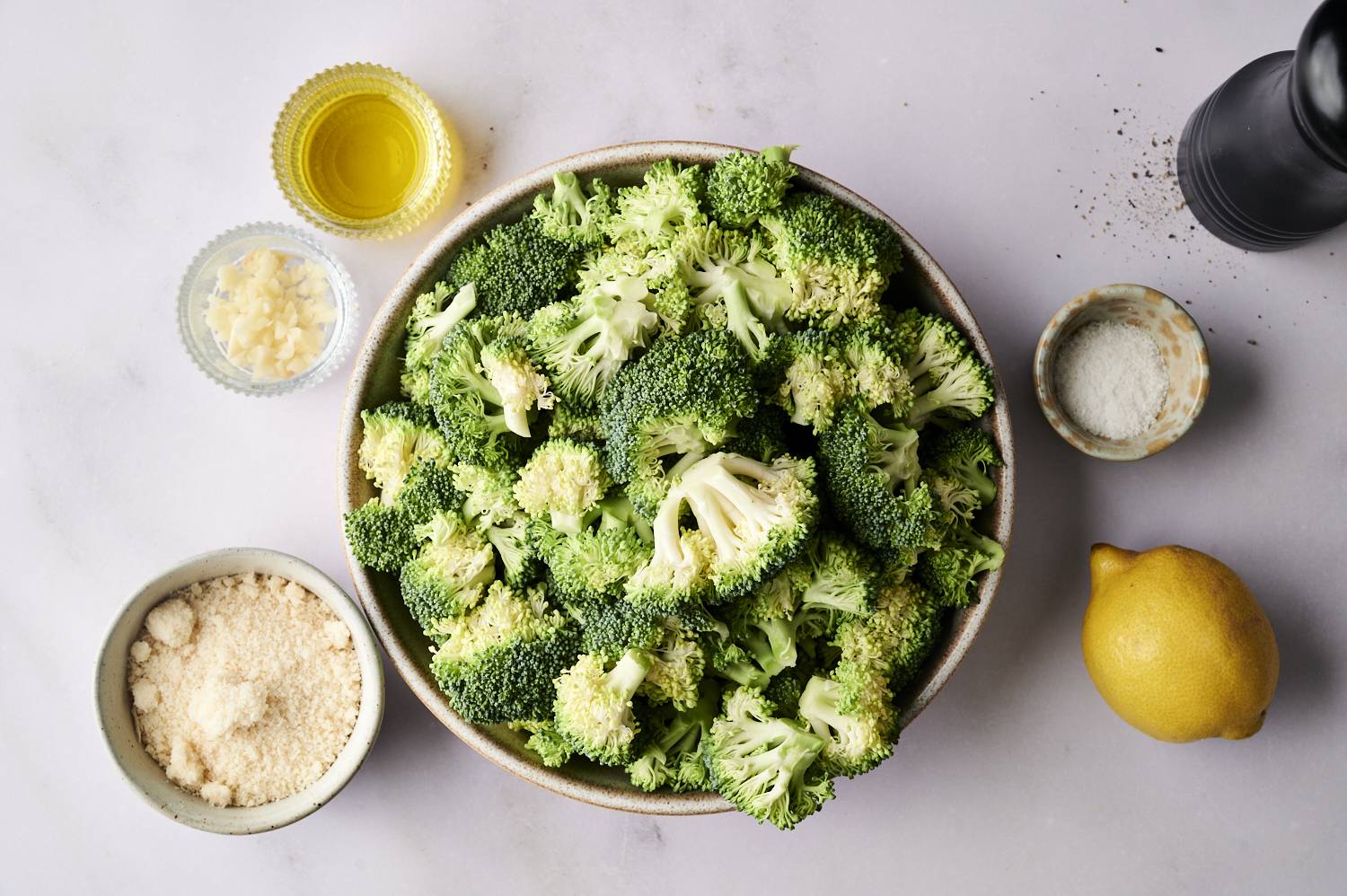 A bowl of fresh broccoli surrounded by garlic, olive oil, lemon, salt, and breadcrumbs on a light countertop.