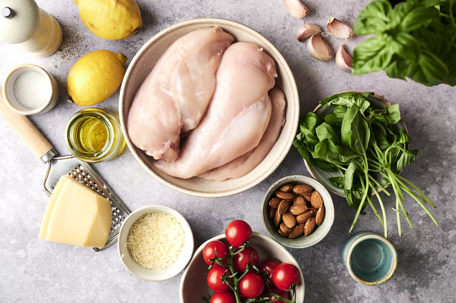 Fresh ingredients for pesto chicken, including raw chicken breast, basil, cherry tomatoes, almonds, garlic, parmesan cheese, olive oil and lemon, are laid out on the countertop.