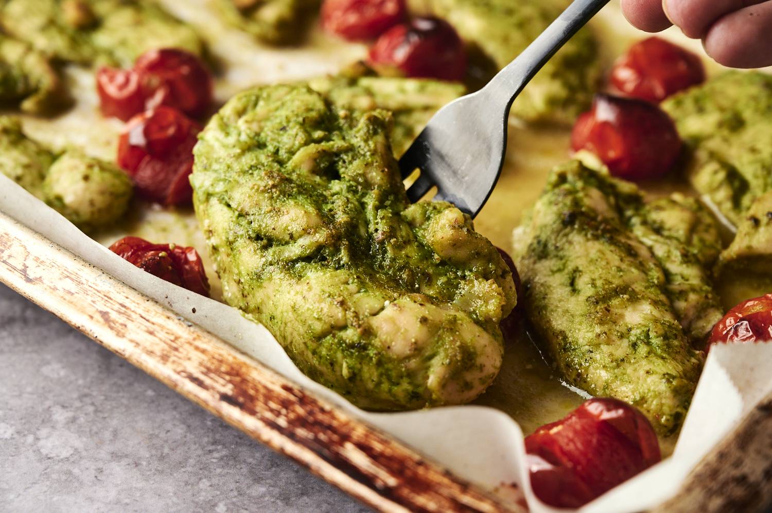 Close-up of grilled chicken slathered in vibrant green pesto sauce and served with roasted cherry tomatoes on a parchment-lined baking sheet.