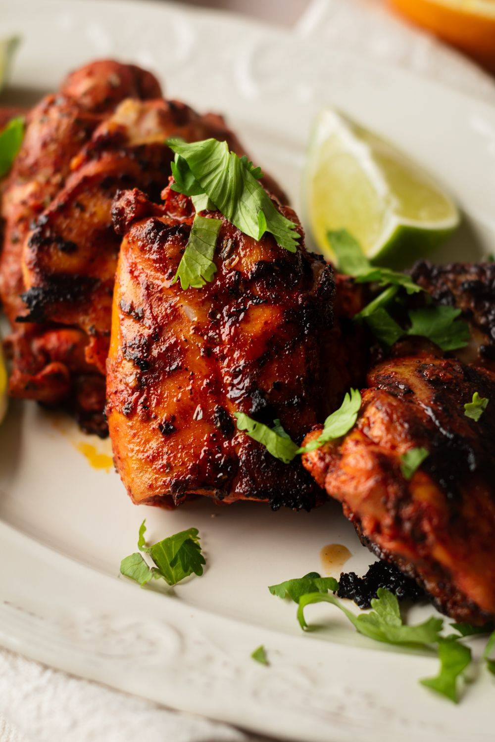 Grilled chicken pieces garnished with fresh cilantro and lime wedges, served on a decorative white plate.