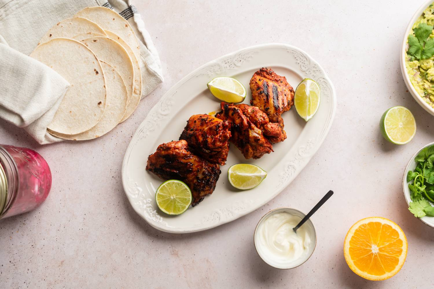 Grilled chicken thighs with lime wedges on a white platter, accompanied by tortillas, guacamole, and sour cream in a cozy setting.