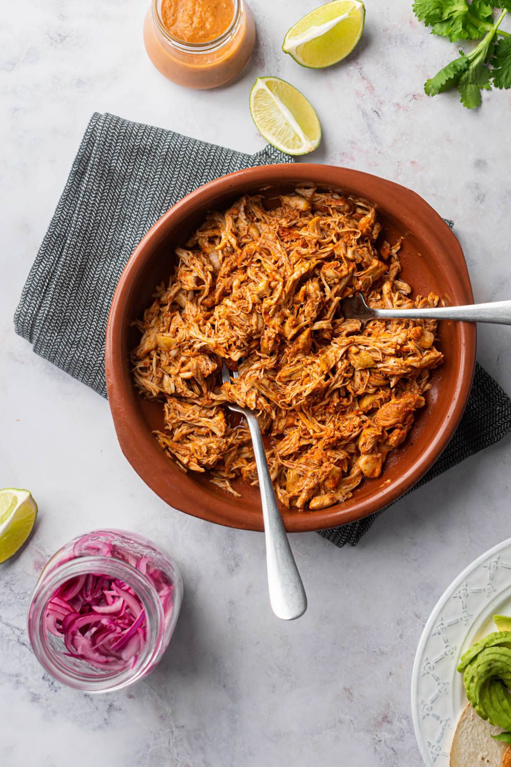Shredded pollo pibil in citrus juices and achiote in a bowl with limes, cilantro, salsa, and pickled onions on the side.