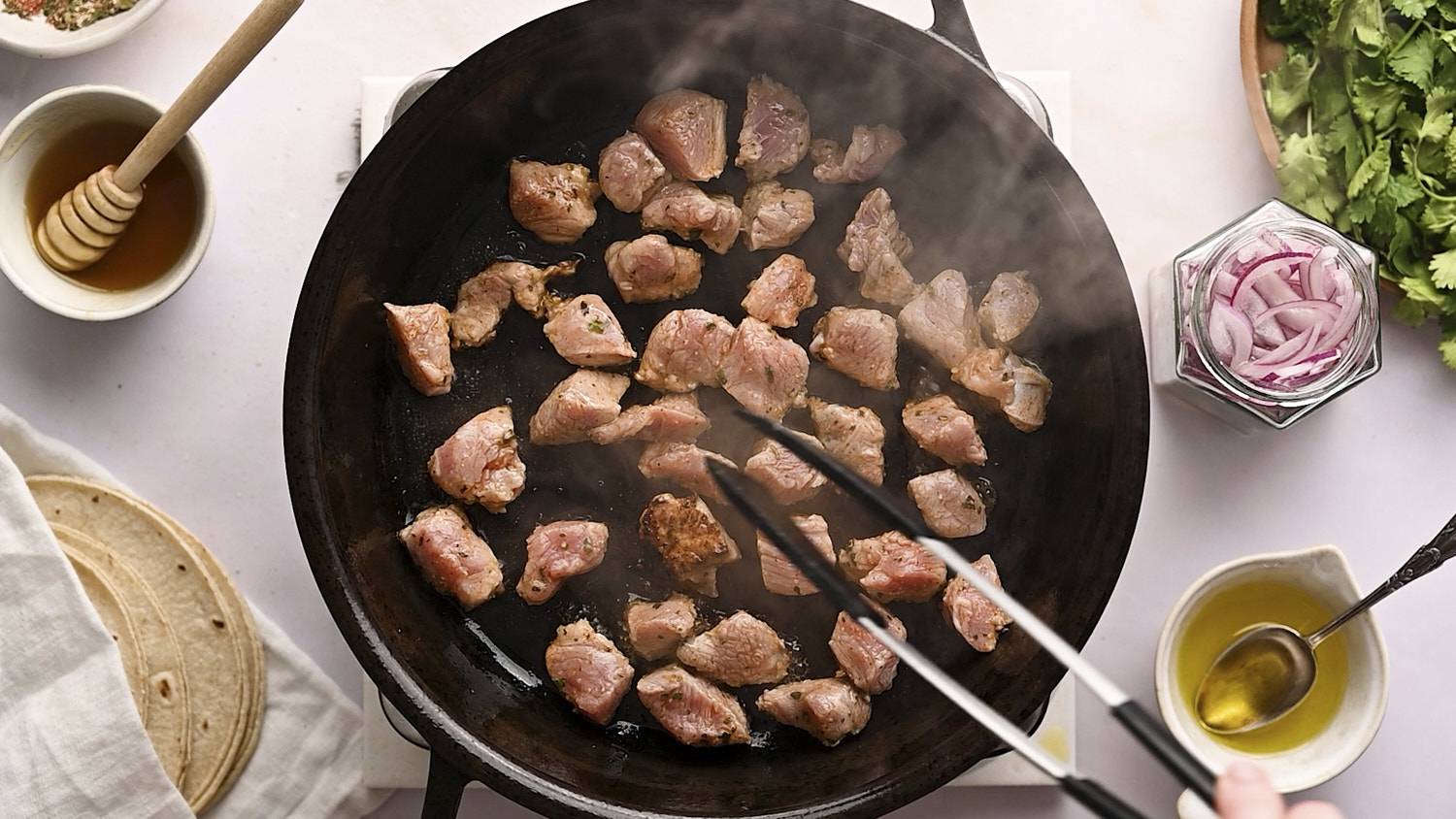 Pieces of pork tenderloin in a cast iron skillet being flipped by tongs.