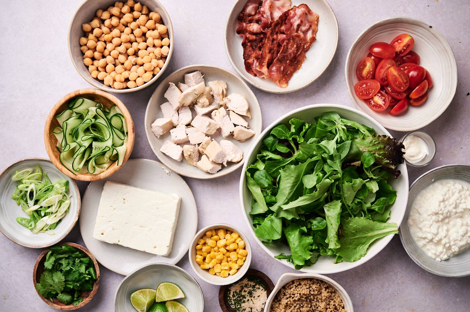 Ingredients for protein packed salad bowls including greens, chicken, chickpeas, cheese, bacon, veggies, and dressing.