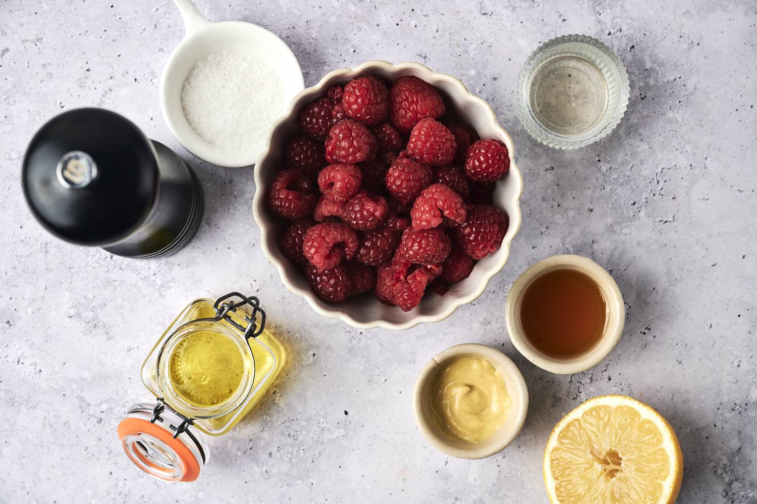 A bowl of fresh raspberries surrounded by olive oil, honey, mustard, salt, lemon, and vinegar on a light textured surface.