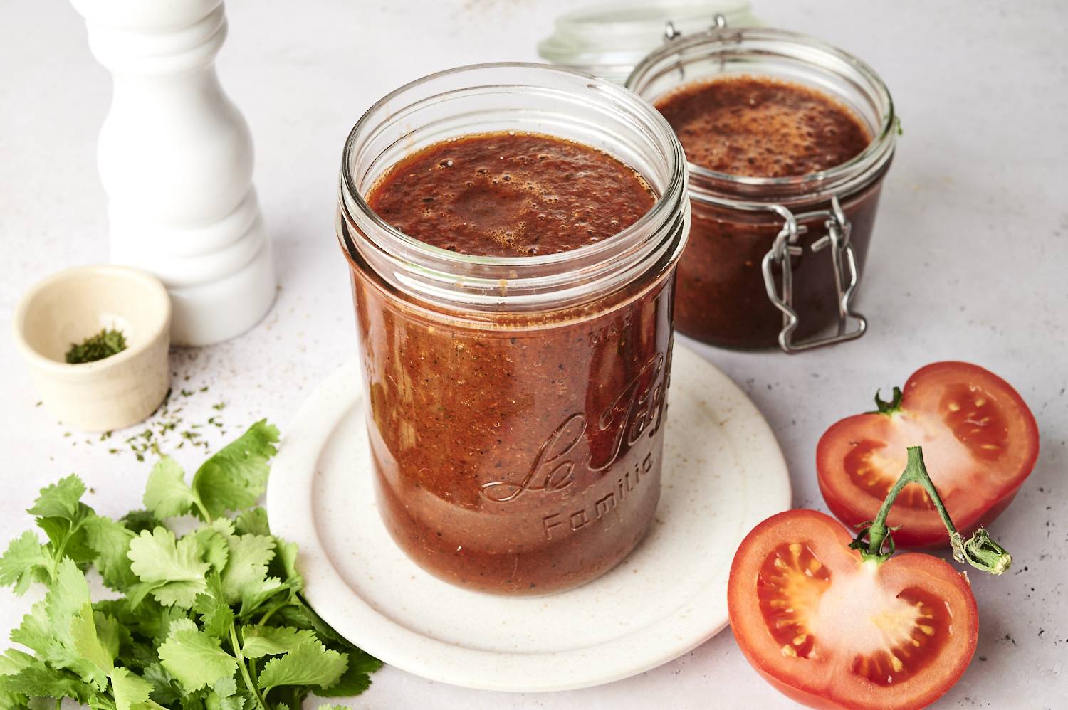 A glass jar filled with vibrant red enchilada sauce, surrounded by fresh cilantro leaves, a halved tomato, and a small bowl of dried herbs, set on a white plate and light background.