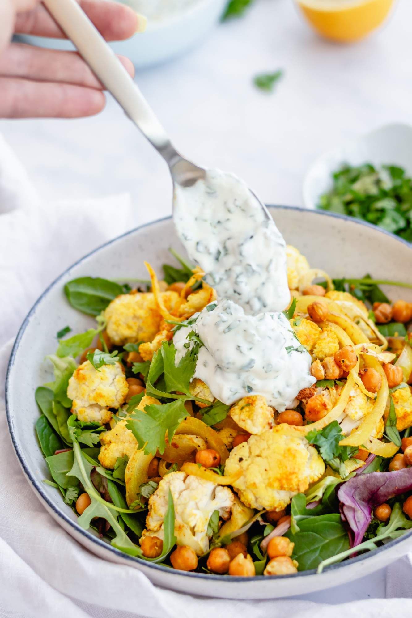 Roasted chickpeas and cauliflowers with onions and greens in a bowl with yogurt sauce being spooned on top.