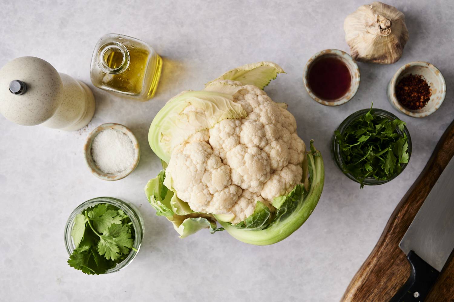 Whole head of cauliflower surrounded by fresh herbs, garlic, olive oil, salt, and seasonings on a gray surface.
