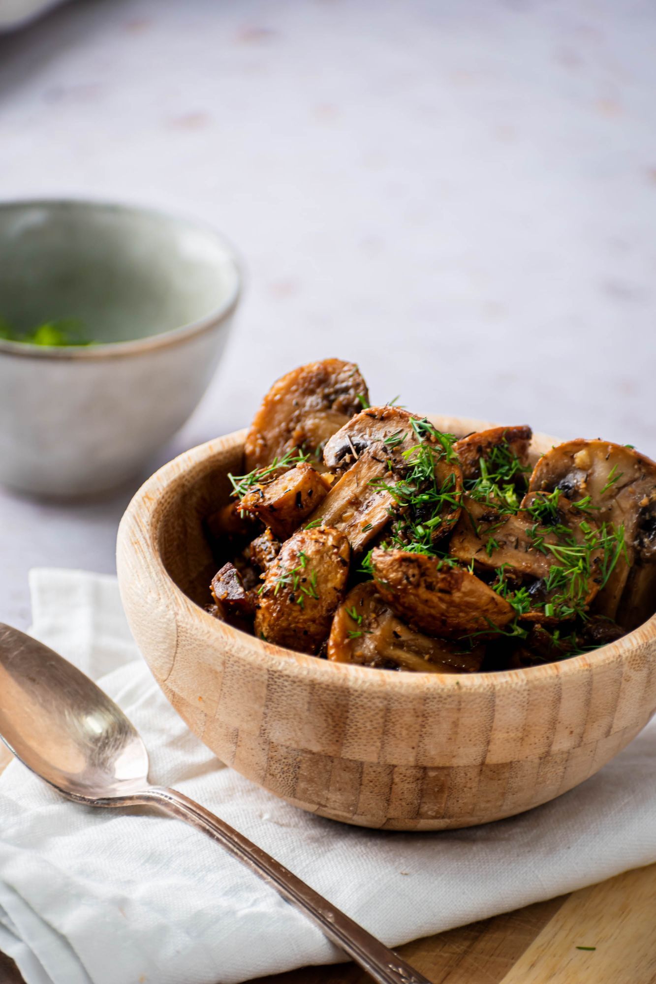 Garlic roasted mushrooms in a wooden bowl with a spoon and herbs on the side.