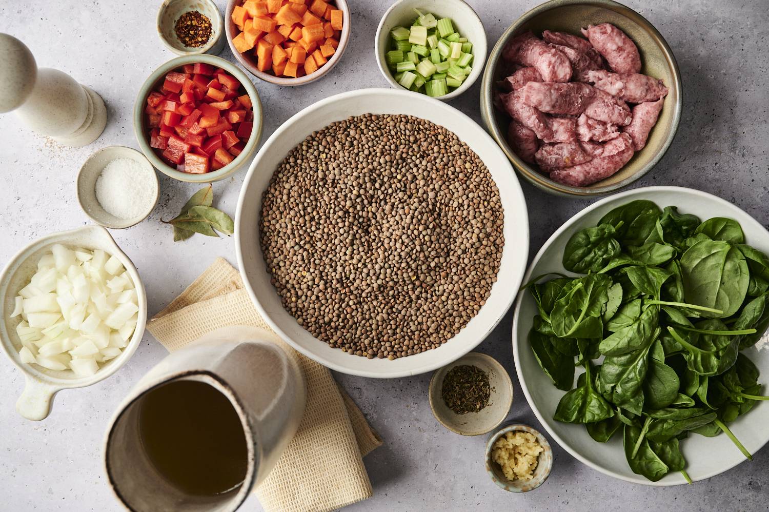Overhead view of sausage lentil soup ingredients arranged in bowls, including dry lentils, fresh spinach, sausage, chopped onions, carrots, celery, red bell peppers, garlic, bay leaf, spices, Parmesan, and broth.