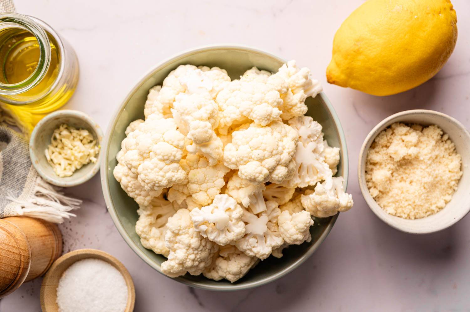 Bowl of cauliflower next to a lemon, a jar of olive oil, minced garlic, a bowl of grated cheese, salt, and pepper mill on a light marble surface.