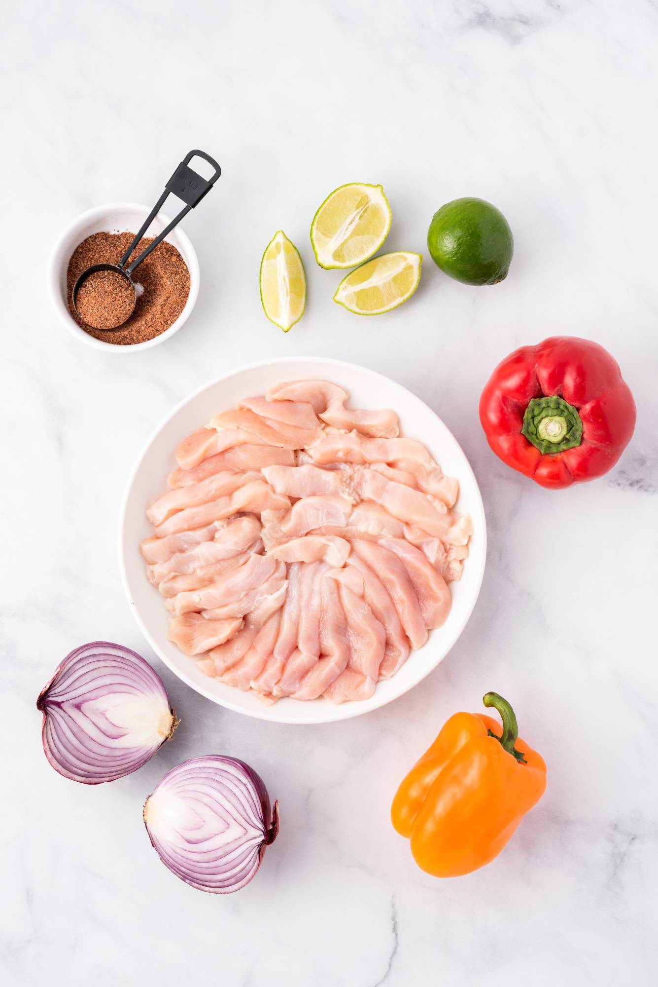 Ingredients for sheet pan chicken fajitas including sliced chicken breast, bell peppers, lime wedges, red onion, and fajita seasoning.