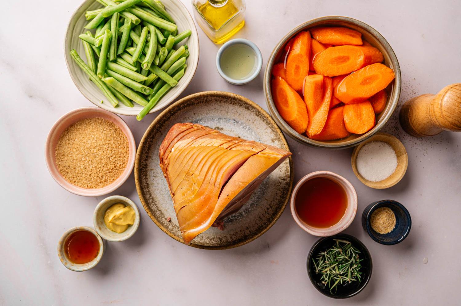 Ingredients for a roast lie on a marble surface: a seasoned ham centerpiece, green beans, carrot slices, mustard, honey, salt, pepper, olive oil, brown sugar, and rosemary.