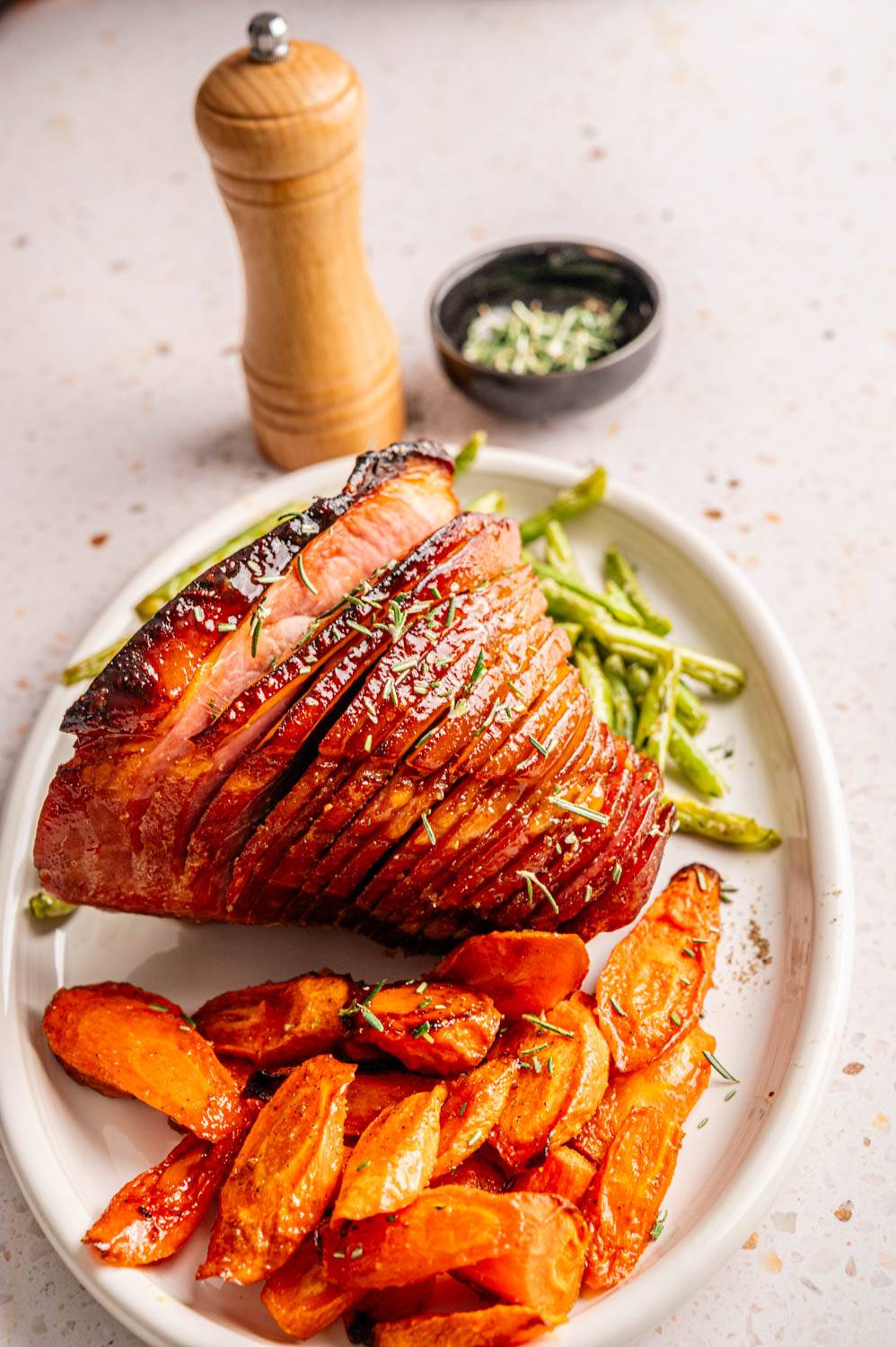 Sliced glazed ham with rosemary on a white plate, accompanied by roasted carrots and green beans. A pepper grinder and herbs in the background.