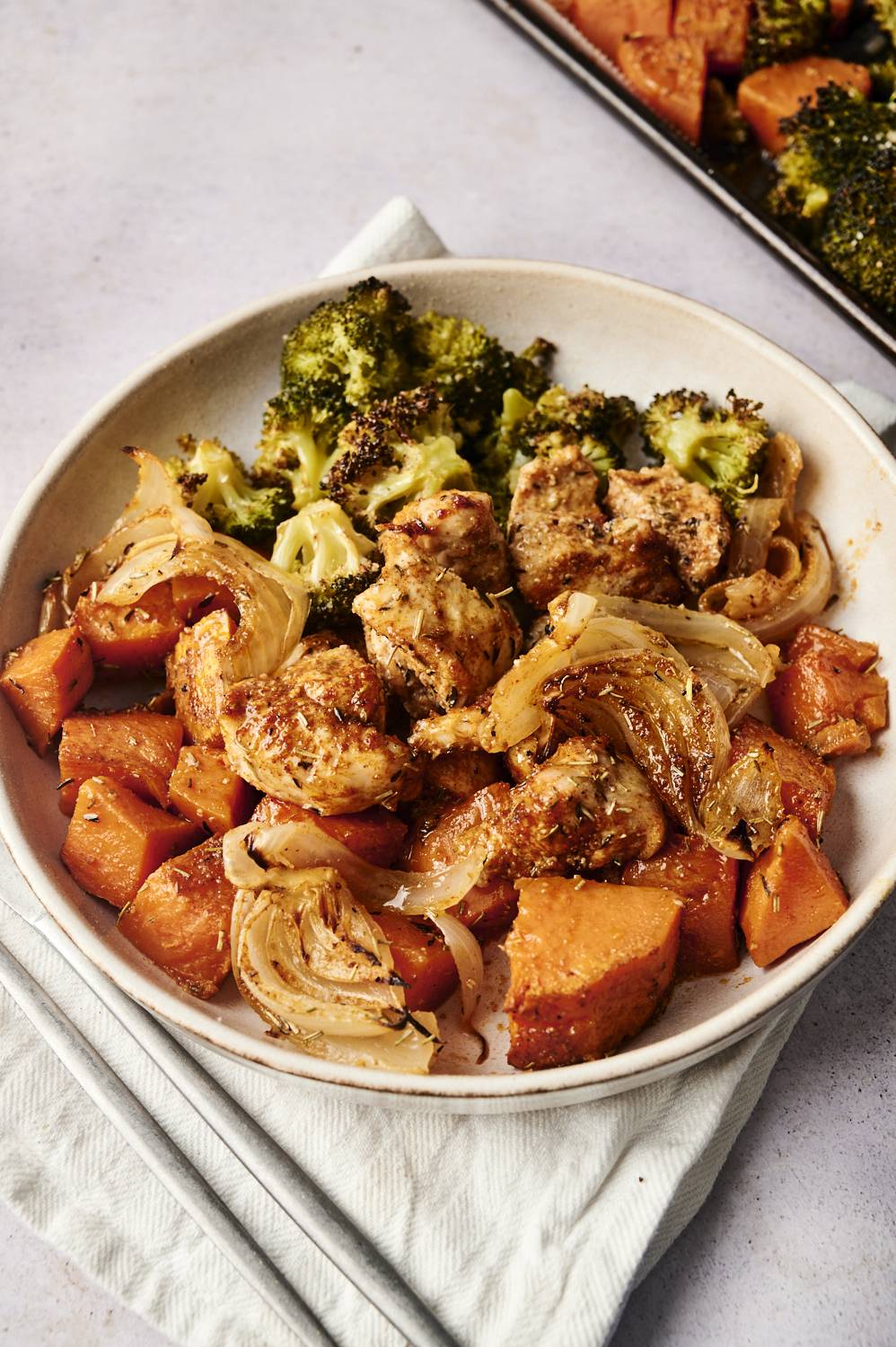 A close-up of a bowl filled with roasted herb-seasoned chicken, caramelized onions, tender sweet potato chunks, and roasted broccoli. The dish is served on a light fabric napkin with a portion of the sheet pan meal visible in the background.